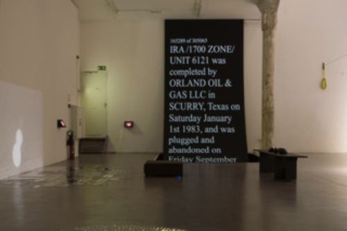 An impression of an installation: A room with white walls and an old column in the middle; there's a projection on the left, a beamer in the middle, and a bank on the right. On the background, a big black screen with the words "IRA / 1700 Zone / UNIT 6121 was completed by ORLAND OIL & GAS LLC in SCURRY, Texas on Saturday, January 1st, 1983, and was plugged and abandoned..."